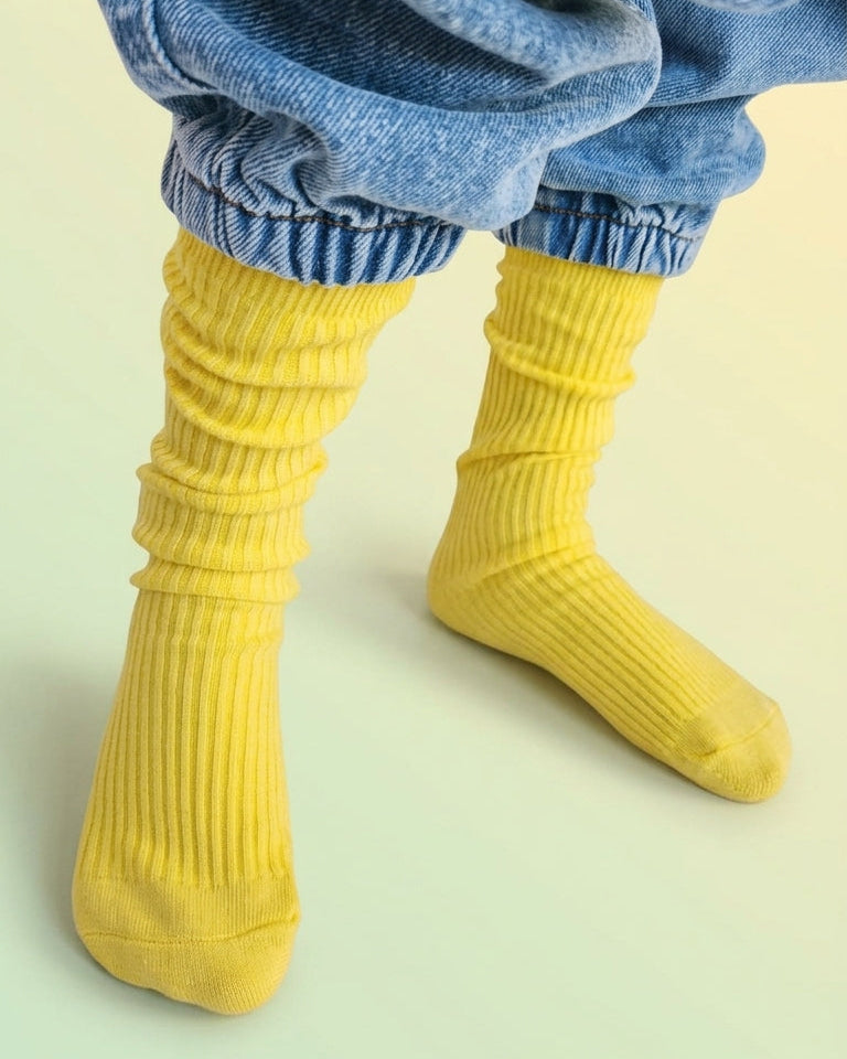 A close-up of a childs's legs wearing bright yellow ribbed slouch socks and light blue baggy jeans, standing against a pale yellow-green background.