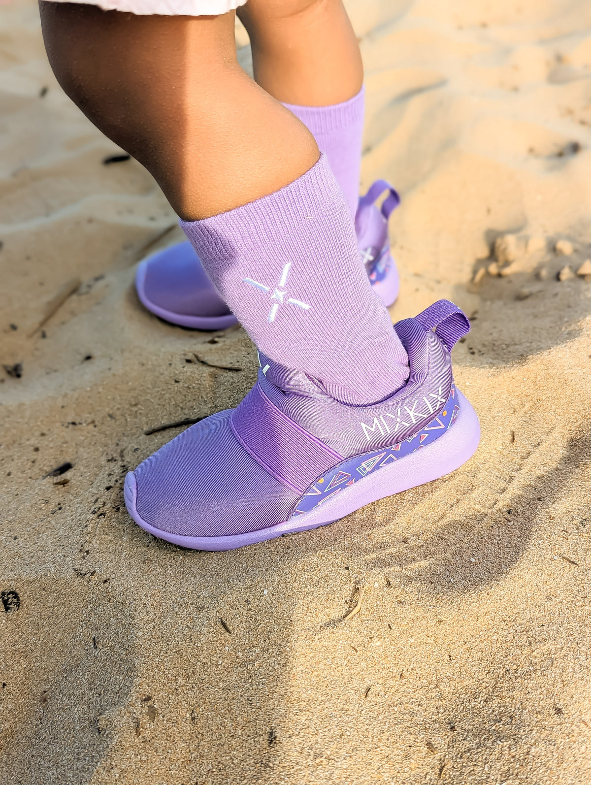 A child wearing lavender-purple socks and matching shoes on a sandy beach. The shoes feature a wide elastic strap for slip-on wear.
