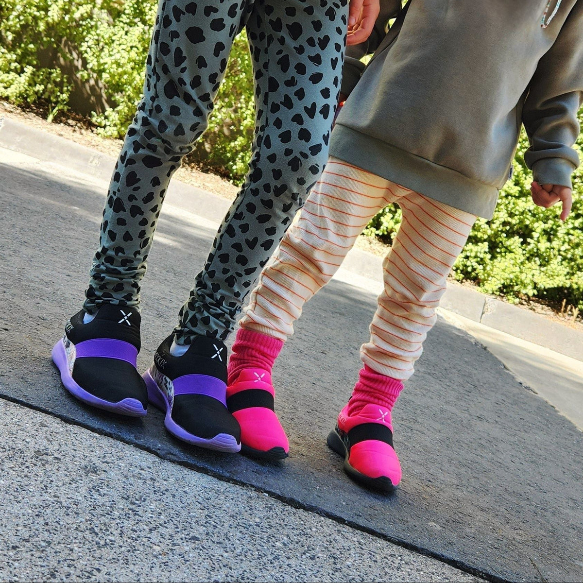 Two children outdoors on a paved walkway wearing and black sneakers with purple soles and pink sneakers with pink socks.
