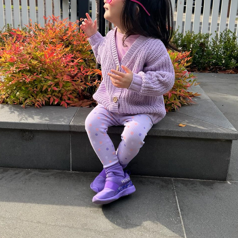 A young child wearing a light purple knit cardigan, matching leggings, and purple sneakers with matching socks. 