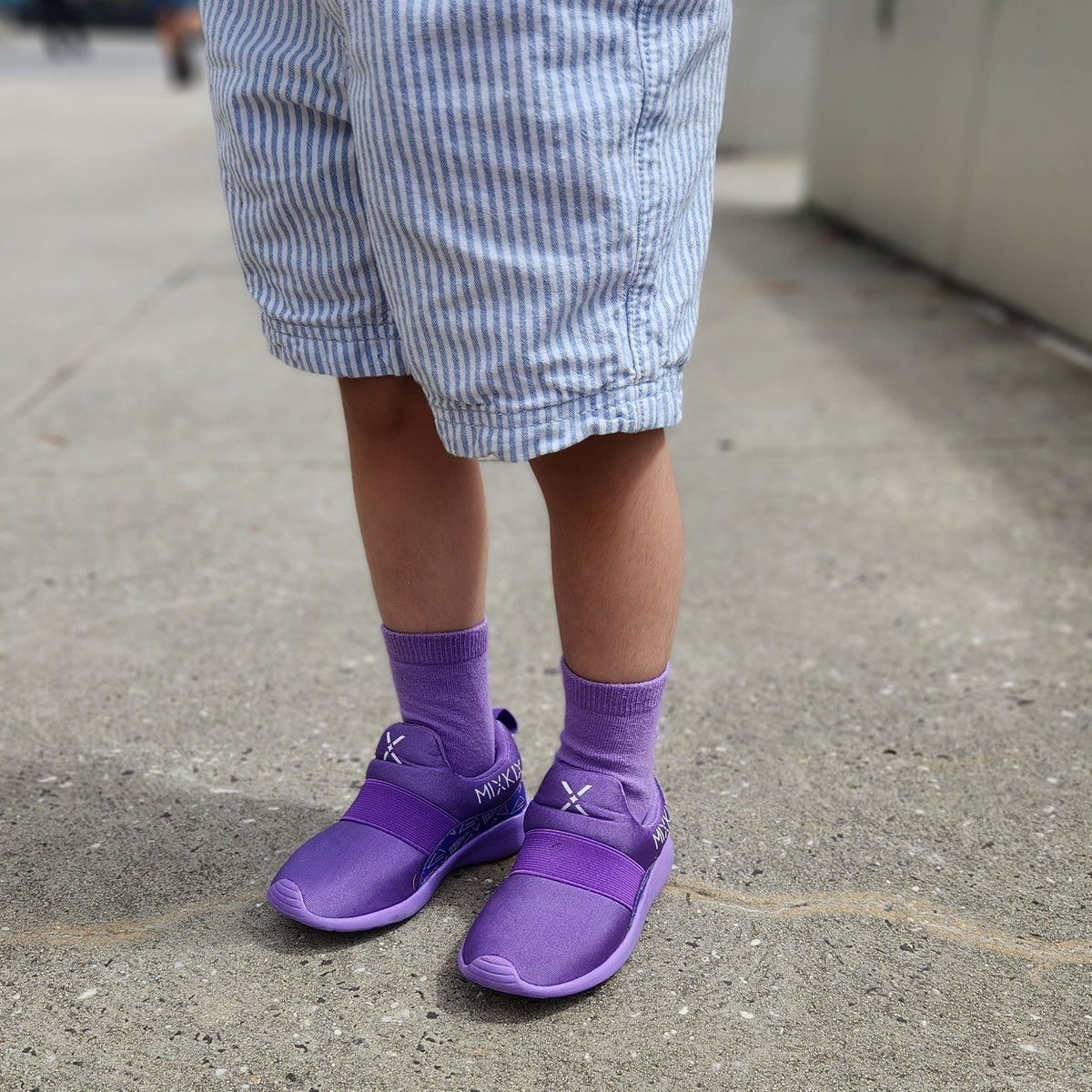 A child wearing purple slip-on shoes and matching purple socks, standing on a concrete outdoor walkway. The child is also wearing blue-and-white vertically striped shorts.