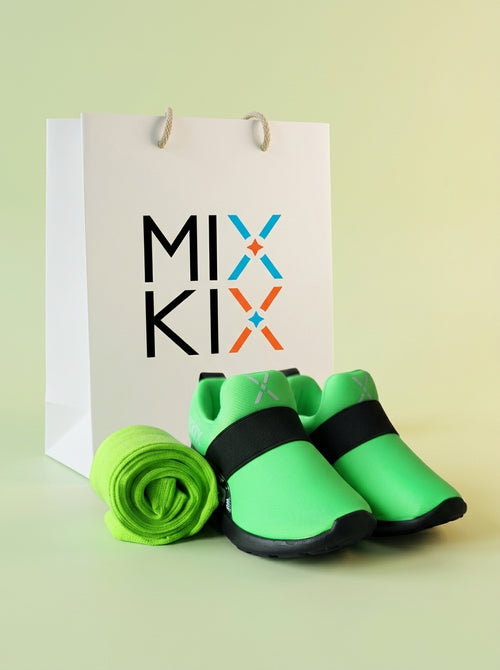 A pair of neon green and black kids shoes with matching rolled-up neon green socks, displayed in front of a white gift bag printed with the brand name 'MIX KIX' on a pale yellow-green background.