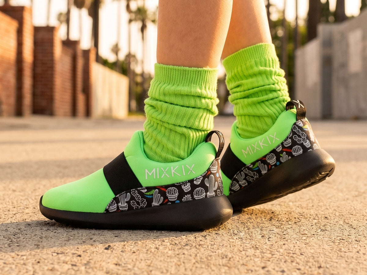 A child wearing bright neon green socks and low-top sneakers with the brand name MIXKIX visible on the side, photographed outdoors on a sunlit paved surface with blurred brick buildings in the background.