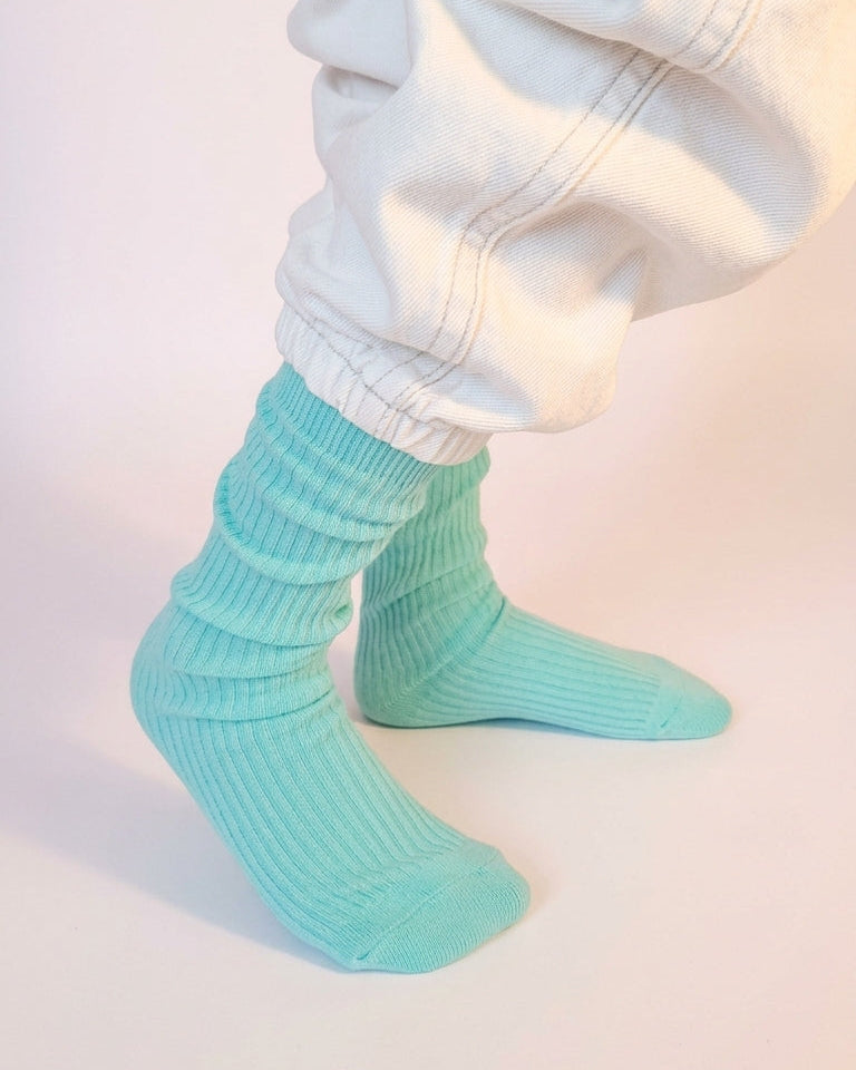 A close-up of a childs feet wearing light Tiffany blue ribbed slouch socks with white baggy jeans, set against a plain light background.