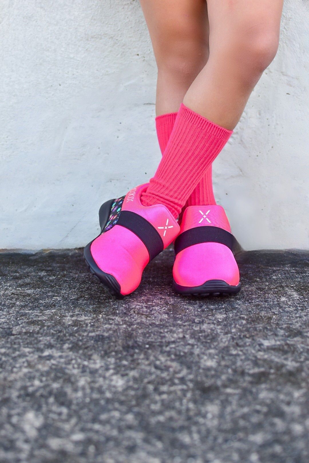 A person wearing bright pink athletic shoes with black straps and matching pink crew socks, standing on a textured gray stone surface against a light gray wall.